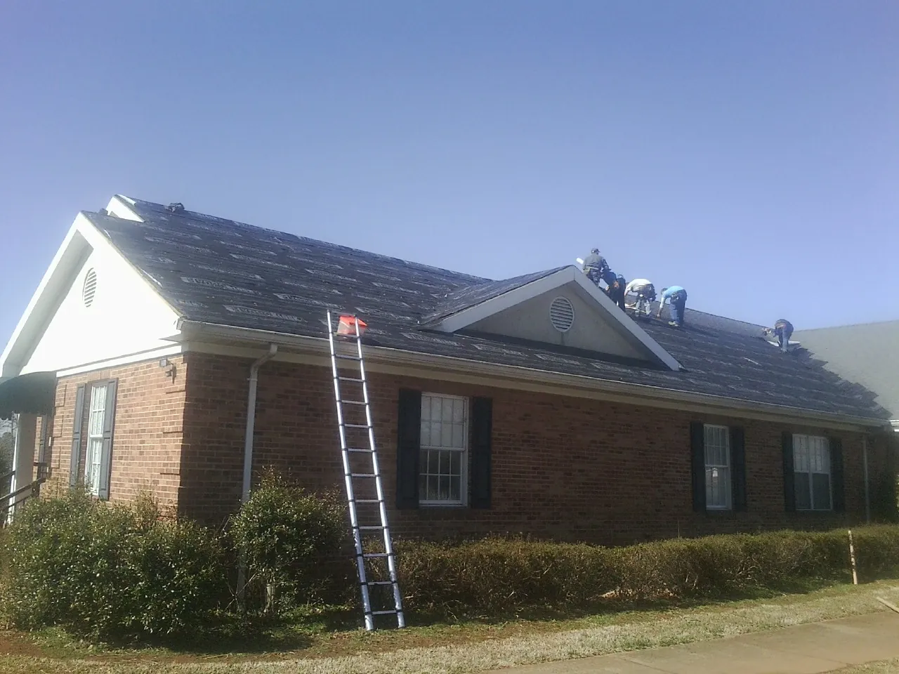Crew tearing shingles off a brick ranch roof with a ladder set — Metal Roof Repair work by our crew in Flint