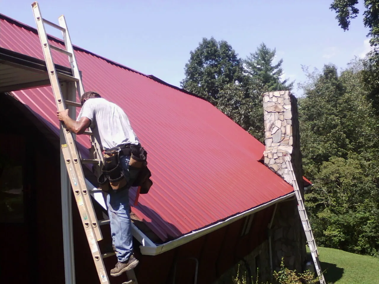 Roofer on a ladder beside a red metal roof and stone chimney — Soffit & Fascia Repair by Roofing Brothers in Flint